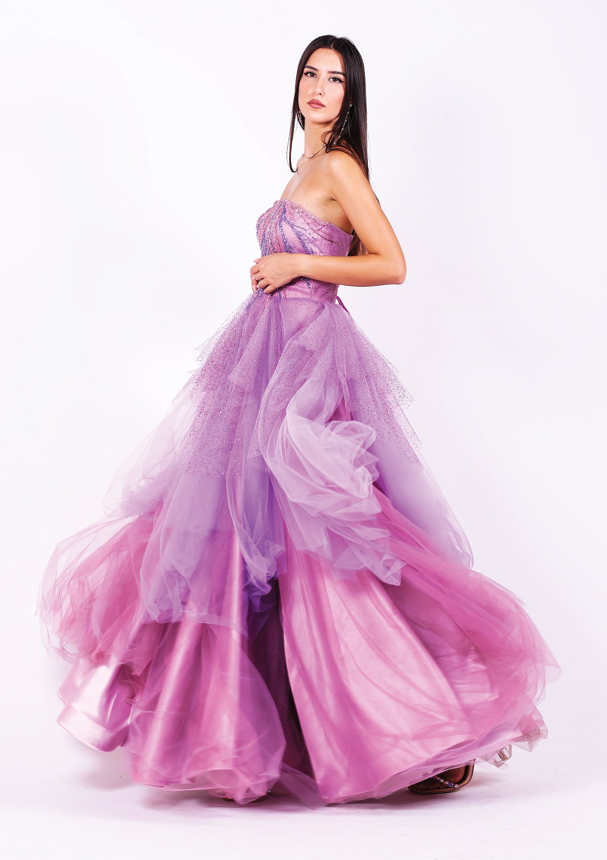 Абитуриентска рокля Ametrine LIZA KAIN София с асиметрични слоеве тюл в различни нюанси лилаво. Корсет с равно деколте и мрежа, покрит с лилава дантела от мъниста. Бухнала пола с волани.