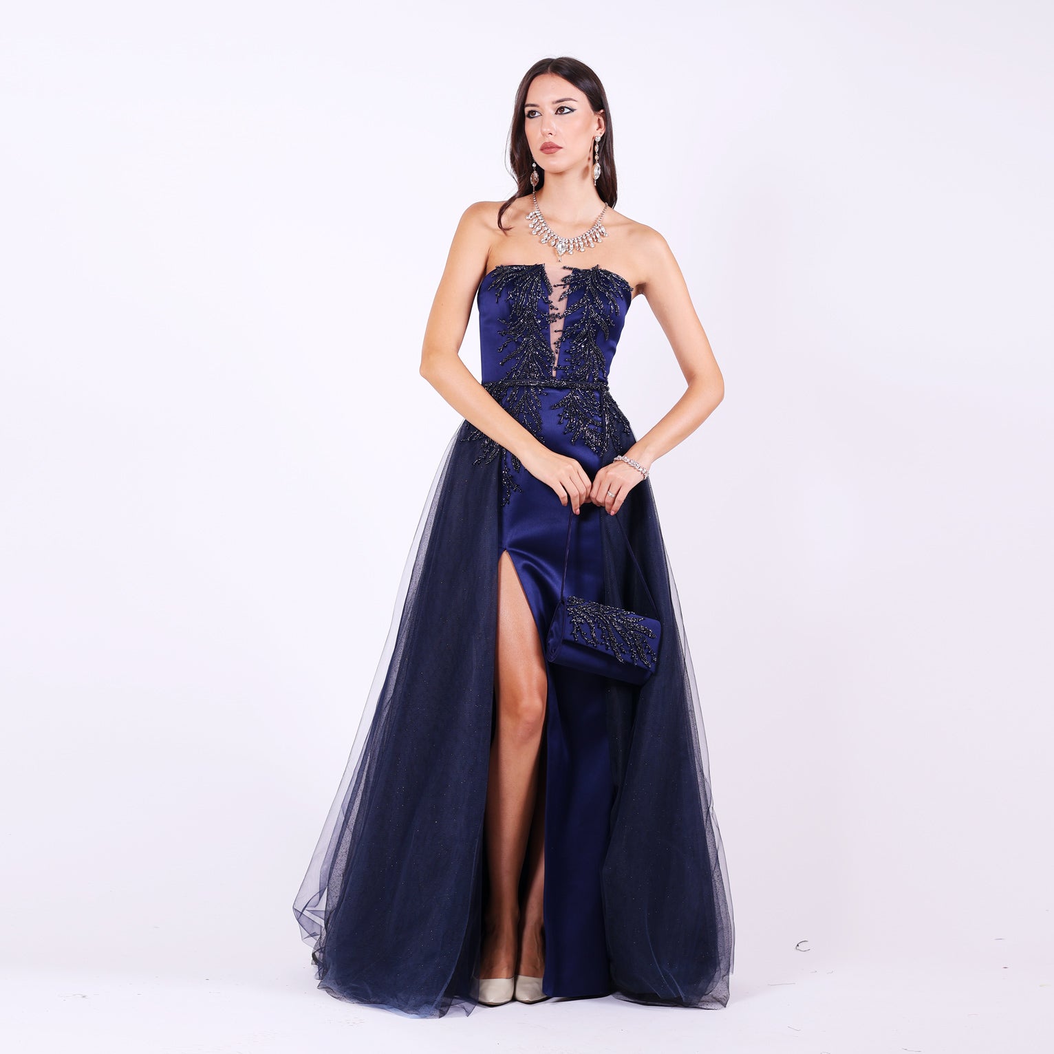 Дълга абитуриентска рокля Mathiola LIZA KAIN София. Официална рокля от тъмносин сатен. Корсет с равно деколте и мрежа, покрит с графитена дантела от камъчета и мъниста. Сатенена пола по тялото с цепка и тюлена пола с колан. Официална чантичка с дръжка.