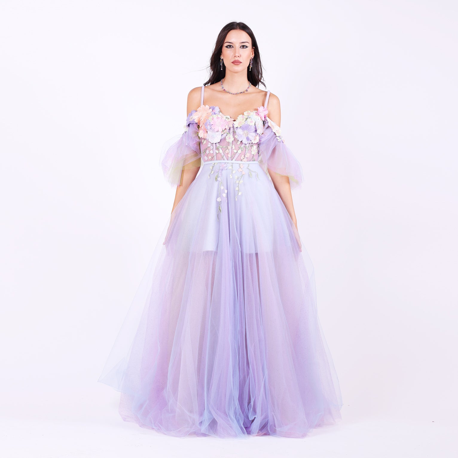 Дълга абитуриентска рокля Faucaria LIZA KAIN София. Официална рокля от светлолилав тюл. Корсет със сърцевидно деколте с тънки презрамки и паднали ръкави.  Апликации от разноцветни цветя в различни размери по горната част. Бухнала пола от различни слоеве тюл с къса сатенена подплата.