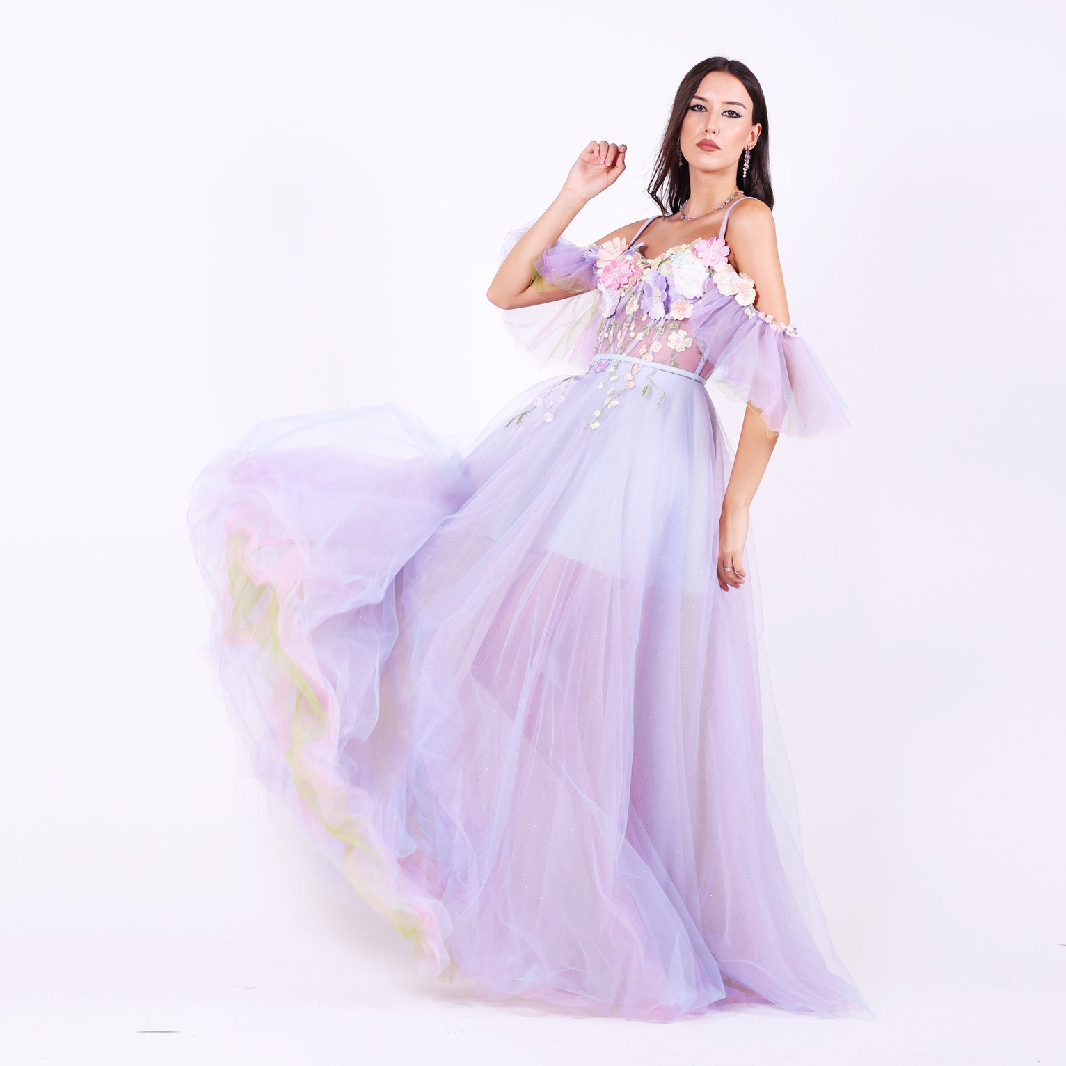 Дълга абитуриентска рокля Faucaria LIZA KAIN София. Официална рокля от светлолилав тюл. Корсет със сърцевидно деколте с тънки презрамки и паднали ръкави.  Апликации от разноцветни цветя по горната част. Бухнала пола от множество слоеве тюл с къса сатенена подплата.