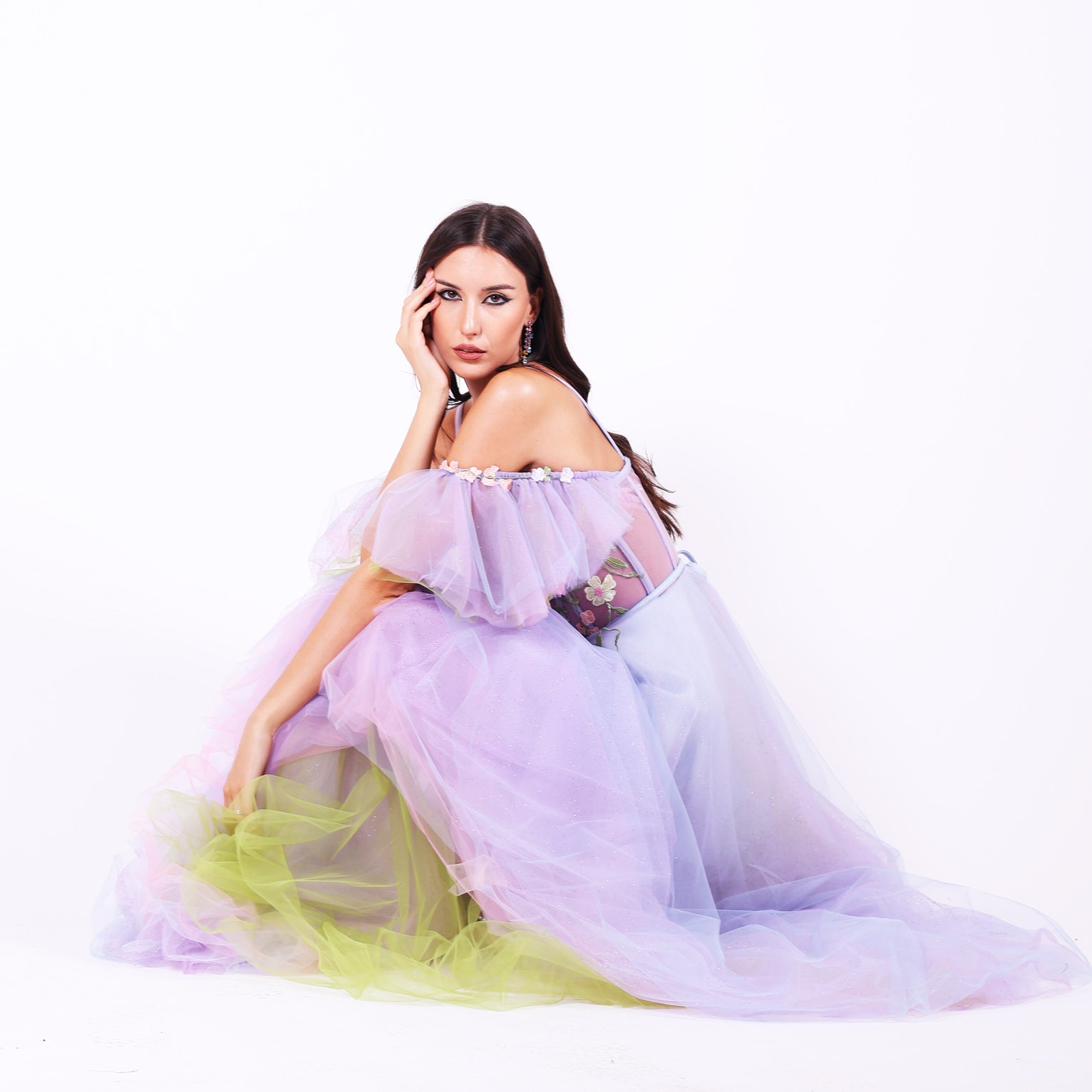 Дълга абитуриентска рокля Faucaria LIZA KAIN София. Официална рокля от светлолилав тюл. Корсет със сърцевидно деколте с тънки презрамки и паднали тюлени ръкави.  Полупрзрачна основа с апликации от разноцветни цветя по горната част. Бухнала пола от множество слоеве тюл с къса сатенена подплата.