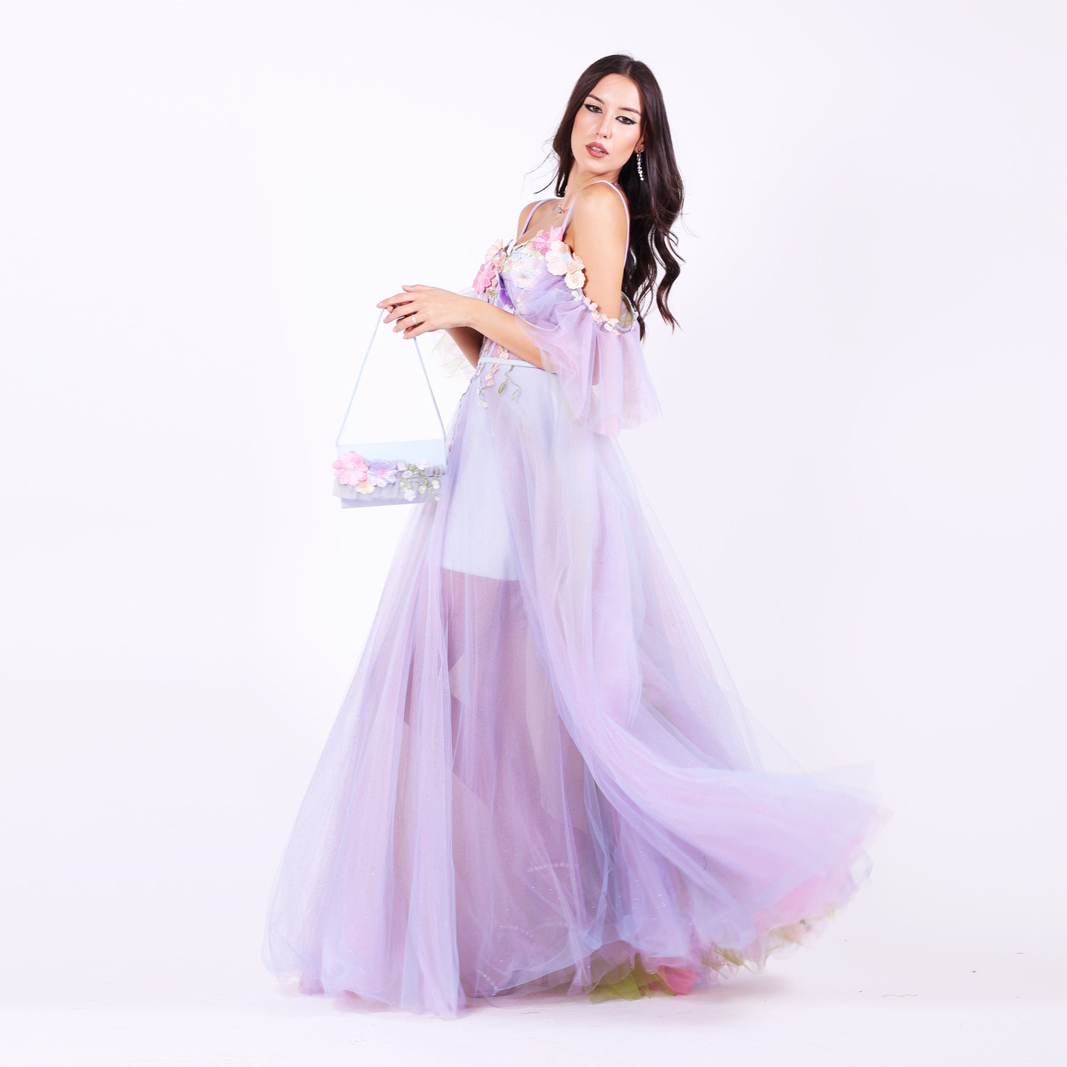 Дълга абитуриентска рокля Faucaria LIZA KAIN София. Официална лилава рокля от тюл. Корсет със сърцевидно деколте с тънки презрамки и паднали ръкави.  Апликации от разноцветни памучни цветя в различни размери по горната част. Бухнала пола от множество слоеве тюл с къса сатенена подплата. Чантичка.