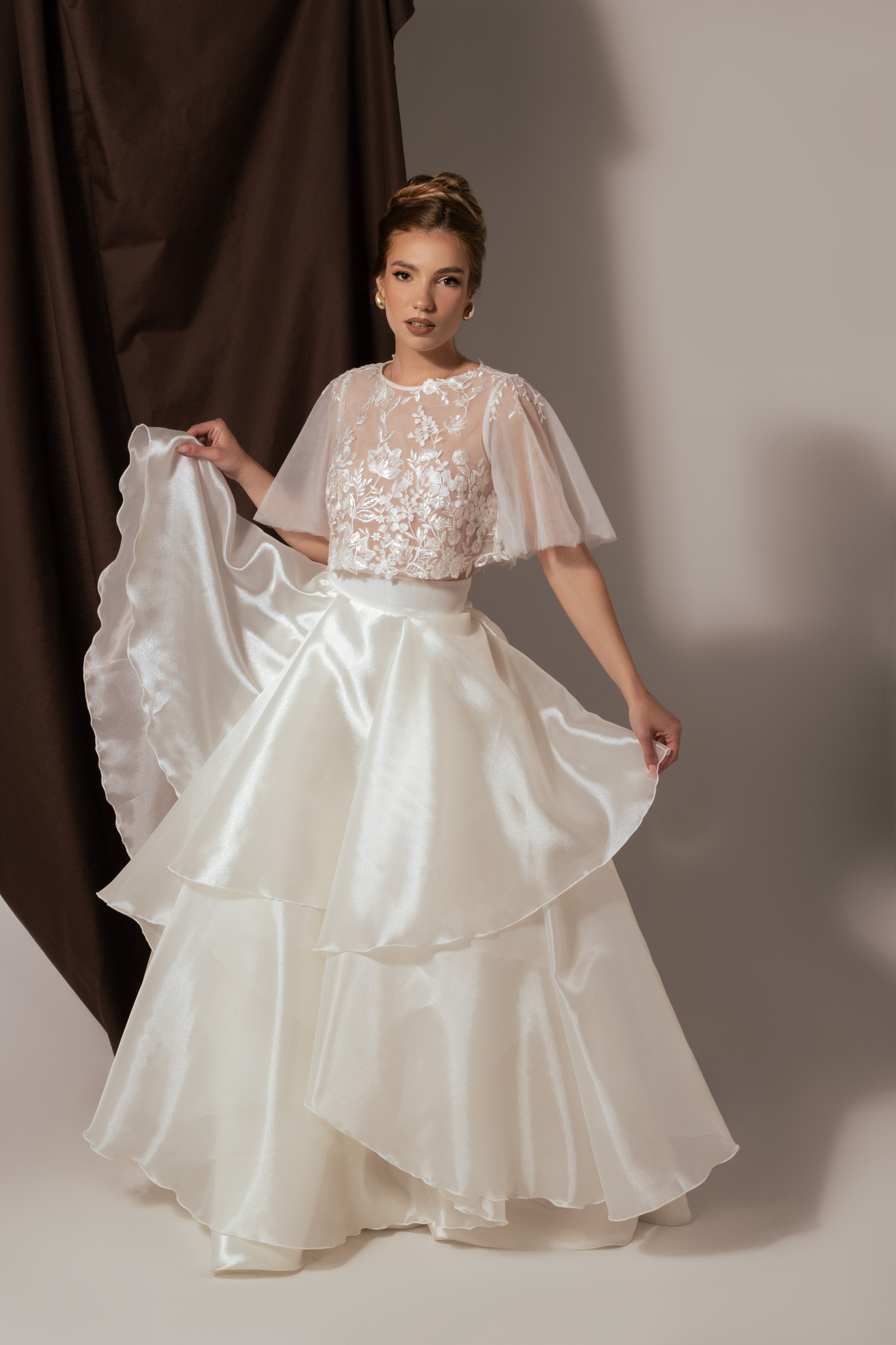 Дълга булчинска рокля Eleanor LIZA KAIN София от две части. Официална бяла рокля. Свободна горна част с ръкави от тюл. Прозрачна основа, покрита с флорална дантела. Обемна пола от органза с колан и воали. 