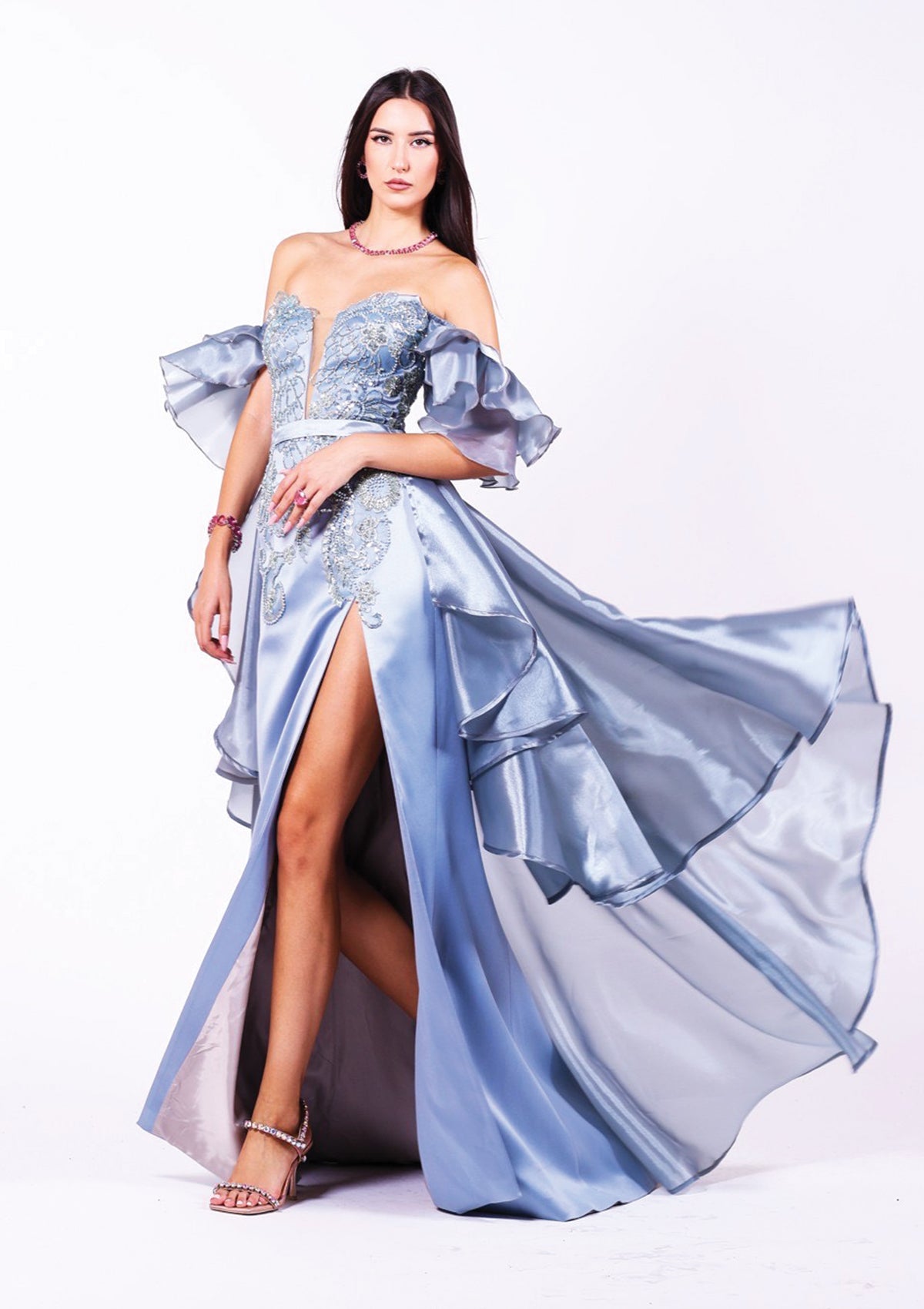 Дълга абитуриентска рокля Moonstone LIZA KAIN София. Официална рокля от светлосин сатен и органза. Корсет с мрежа и паднали къдрави ръкави. Покрита със сребриста флорална дантела от мъниста и перли. Пола с добавен шлейф от органза с колан.